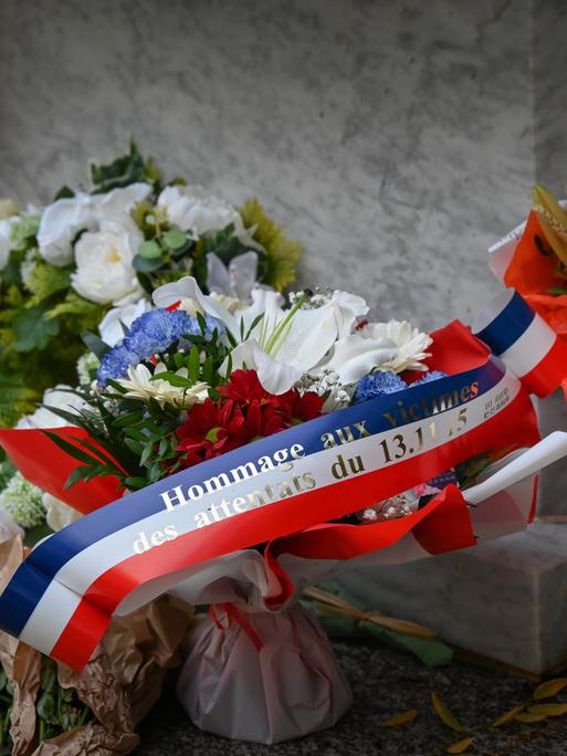 Ein Blumengesteck mit einer Schleife in den Farben der französischen Nationalflagge, auf dem steht: "Hommage an die Opfer der Anschläge vom 13. November 2015". Ein Blumengesteck mit einer Schleife in den Farben der französischen Nationalflagge, auf dem steht: "Hommage an die Opfer der Anschläge vom 13. November 2015".