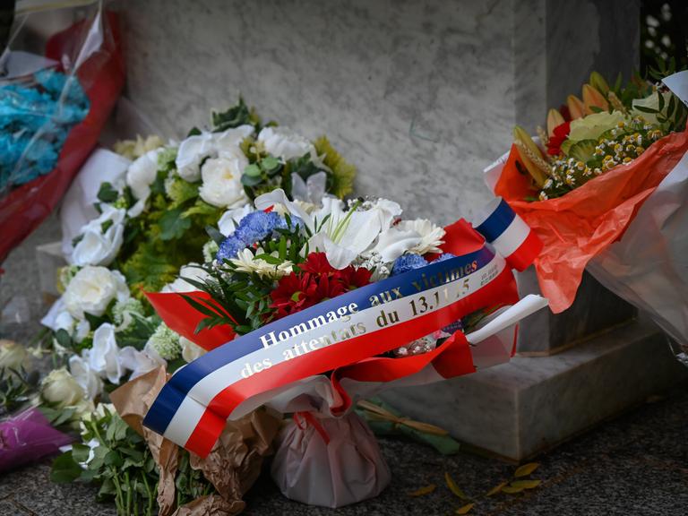 Ein Blumengesteck mit einer Schleife in den Farben der französischen Nationalflagge, auf dem steht: "Hommage an die Opfer der Anschläge vom 13. November 2015". Ein Blumengesteck mit einer Schleife in den Farben der französischen Nationalflagge, auf dem steht: "Hommage an die Opfer der Anschläge vom 13. November 2015".