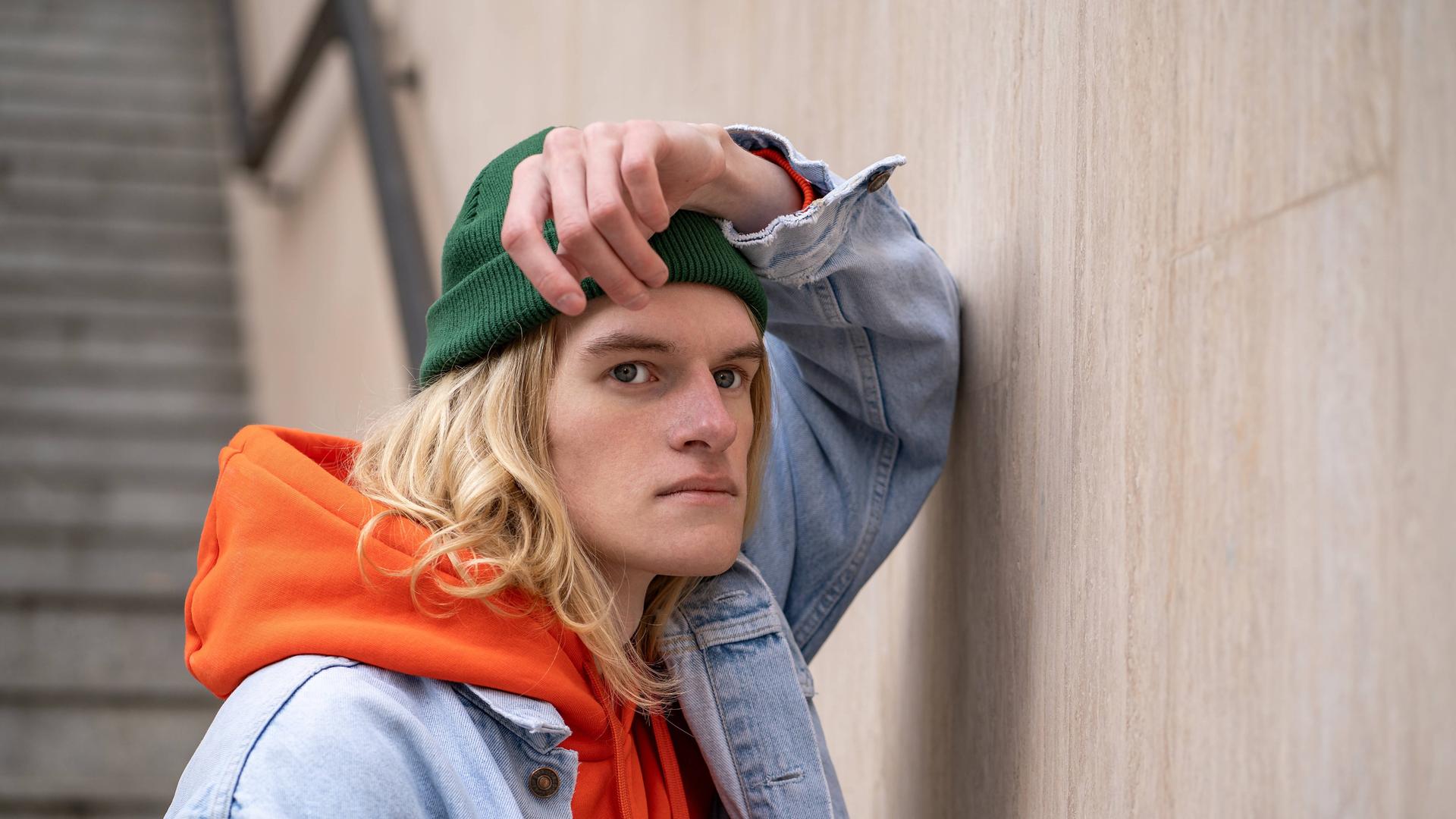 Frustriert schauender Mann mit grüner Mütze, orangen Hoodie und Jeansjacke lehnt an Wand