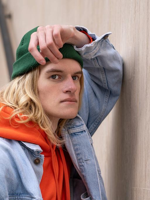 Frustriert schauender Mann mit grüner Mütze, orangen Hoodie und Jeansjacke lehnt an Wand Frustriert schauender Mann mit grüner Mütze, orangen Hoodie und Jeansjacke lehnt an Wand