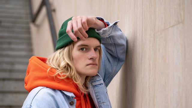 Frustriert schauender Mann mit grüner Mütze, orangen Hoodie und Jeansjacke lehnt an Wand