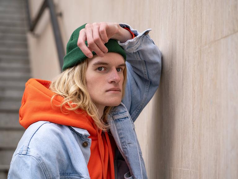 Frustriert schauender Mann mit grüner Mütze, orangen Hoodie und Jeansjacke lehnt an Wand