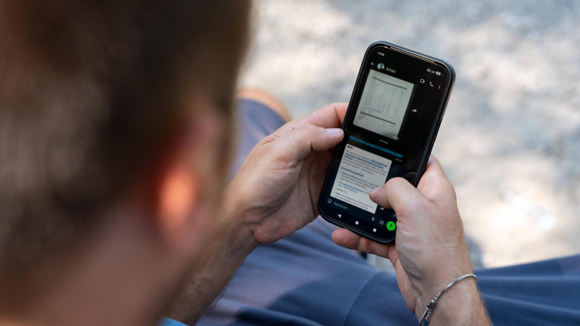 Ein Mann sitzt mit einem Smartphone im Freien und liest auf dem Display.