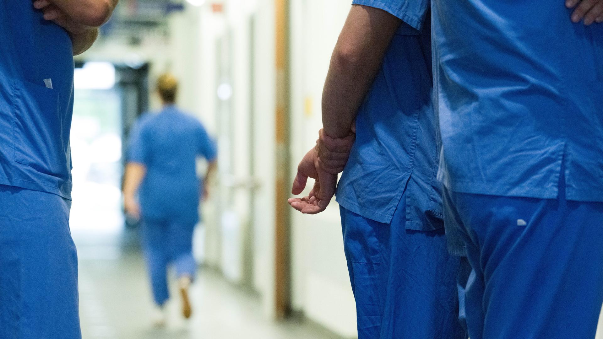Im Gang eines Krankenhauses stehen mehrere medizinische Fachkräfte zusammen. Im Bild sieht man nur ihre Körper, aber nicht ihre Gesichter. Sie tragen blaue Arbeitskleidung. Im Gang eines Krankenhauses stehen mehrere medizinische Fachkräfte zusammen. Im Bild sieht man nur ihre Körper, aber nicht ihre Gesichter. Sie tragen blaue Arbeitskleidung.