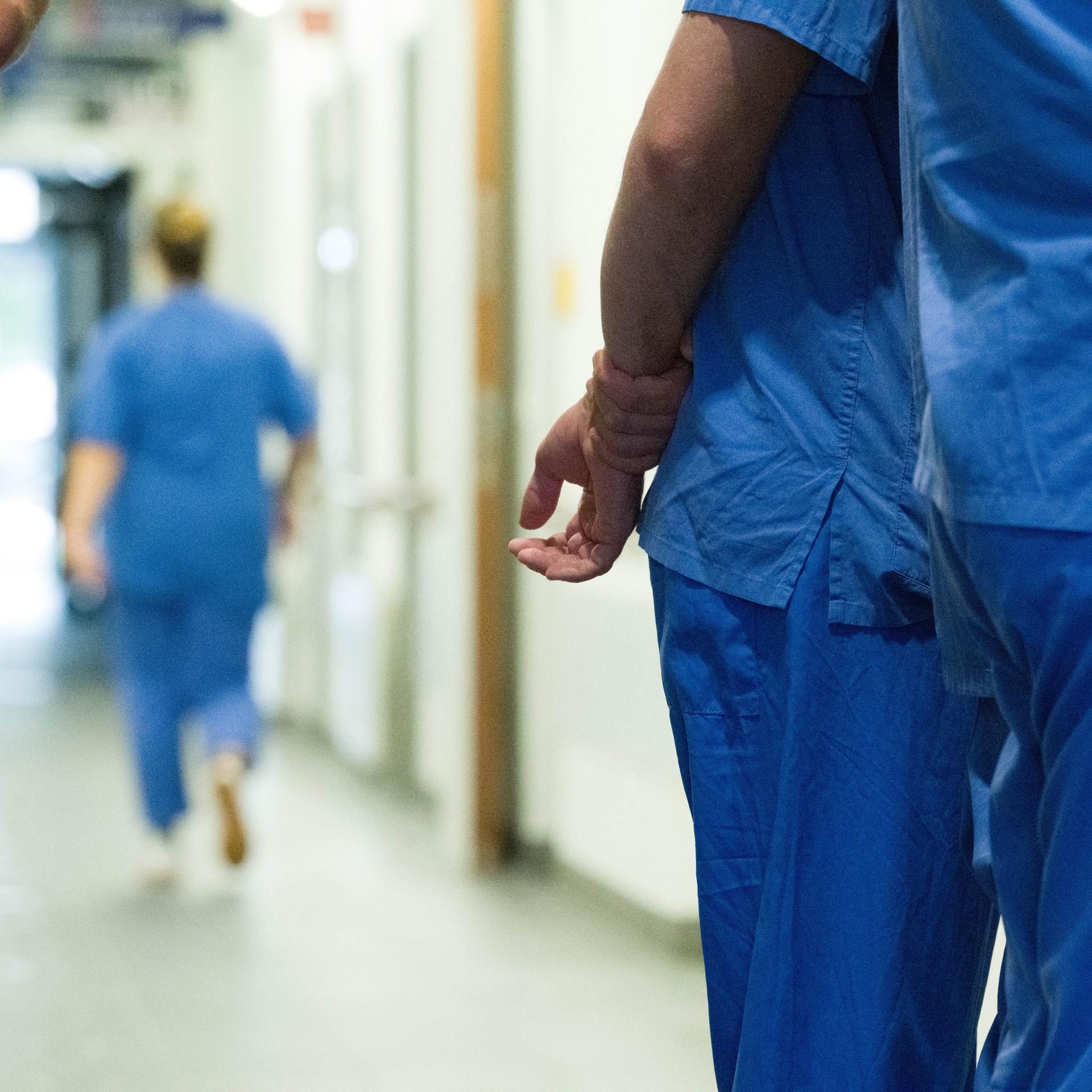 Im Gang eines Krankenhauses stehen mehrere medizinische Fachkräfte zusammen. Im Bild sieht man nur ihre Körper, aber nicht ihre Gesichter. Sie tragen blaue Arbeitskleidung.