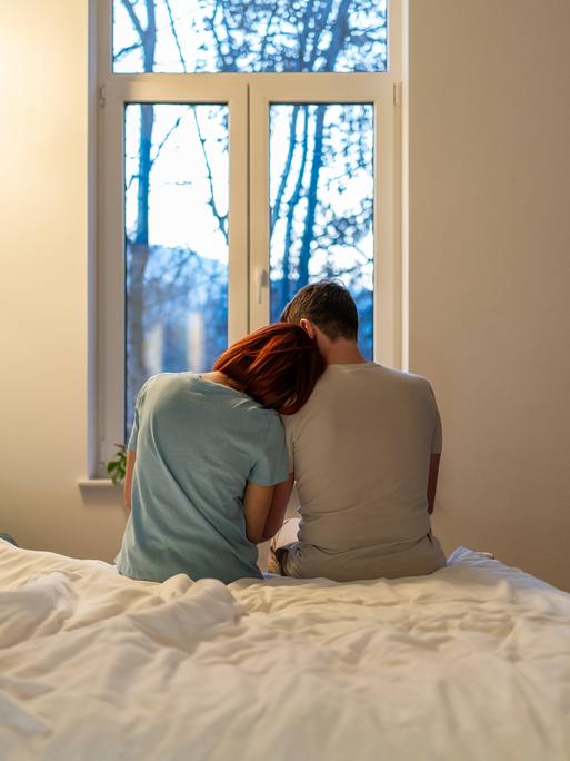 Eine Frau und ein Mann sitzen nebeneinander am Bettrand. Sie lehnt ihren Kopf an seiner Schulter an.