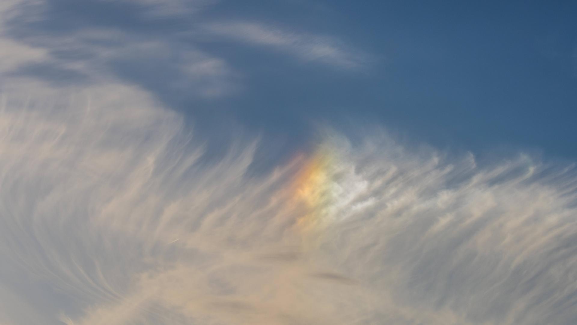 Schleierwolken am blauen Himmel mit einem leuchtenden, regenbogenfarbenen Lichtfleck (Nebensonne).