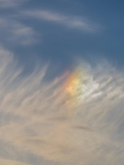 Schleierwolken am blauen Himmel mit einem leuchtenden, regenbogenfarbenen Lichtfleck (Nebensonne).