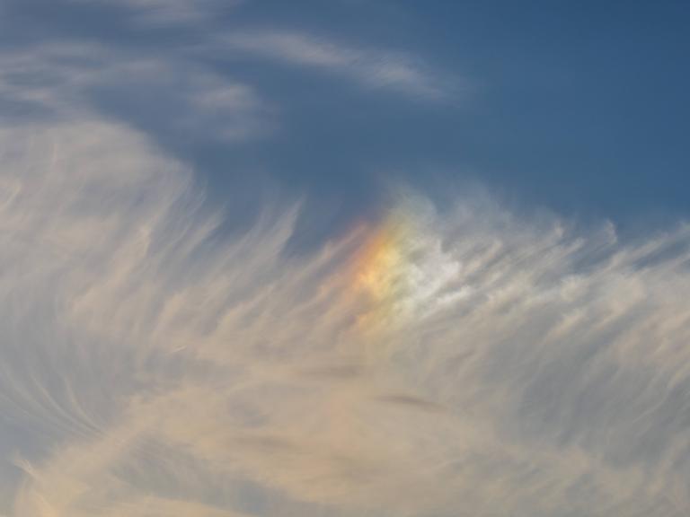 Schleierwolken am blauen Himmel mit einem leuchtenden, regenbogenfarbenen Lichtfleck (Nebensonne).