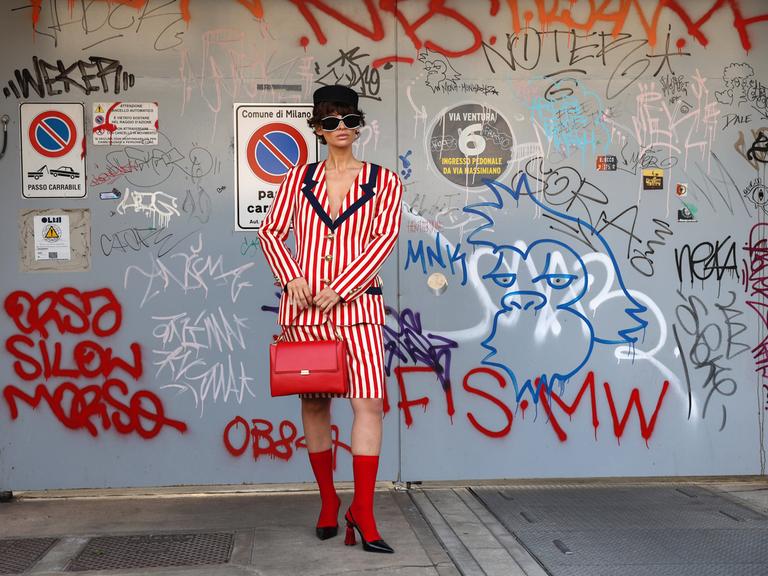 Eine Frau in einem rot-weiß-gestreiften Kostüm, mit roten Socken, roter Handtasche, Hut und Sonnenbrille posiert vor einer mit Grafiti beschmierten Wand.