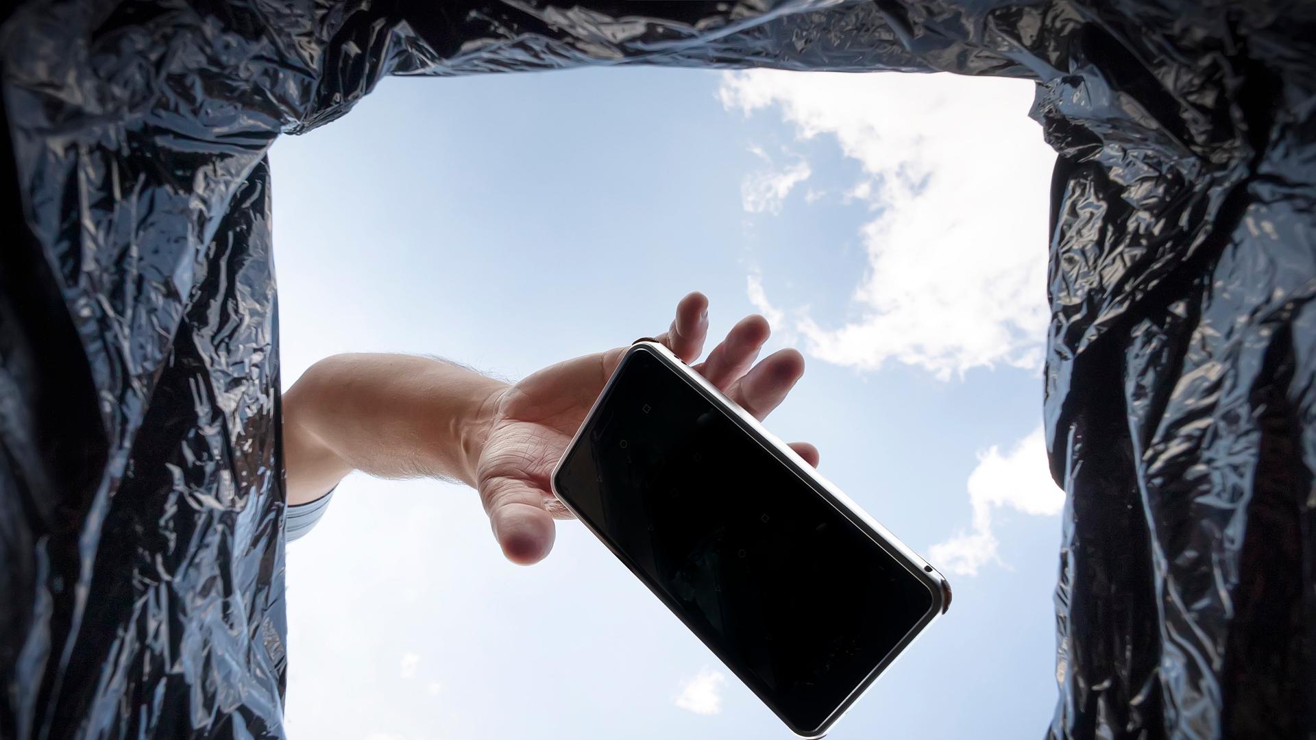 Blick aus einem Mülleimer in den blauen Himmel. Eine Hand wirft ein Smartphone in den Müll.