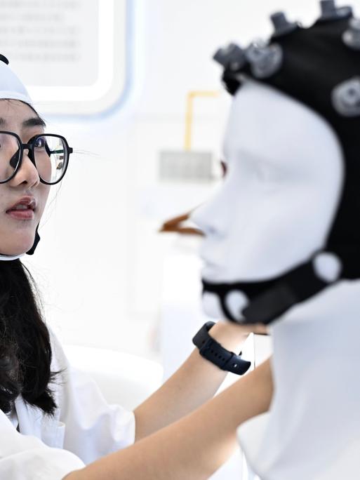 Bei einer Messe in China trägt eine Besucherin eine Art Mütze mit Elektroden, vor ihr steht ein Modellkopf, ebenfalls mit Mütze und Elektroden.