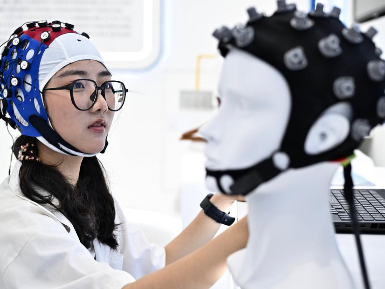 Bei einer Messe in China trägt eine Besucherin eine Art Mütze mit Elektroden, vor ihr steht ein Modellkopf, ebenfalls mit Mütze und Elektroden.