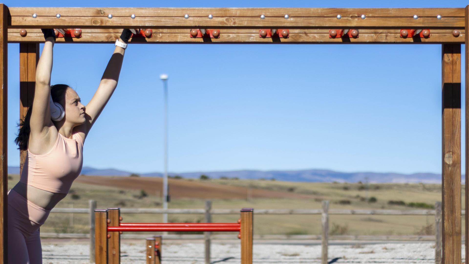 Porträt einer sportlichen Frau beim Klimmzug am Barren an einem sonnigen Tag mit Kopfhörern und Sportkleidung Porträt einer sportlichen Frau beim Klimmzug am Barren an einem sonnigen Tag mit Kopfhörern und Sportkleidung