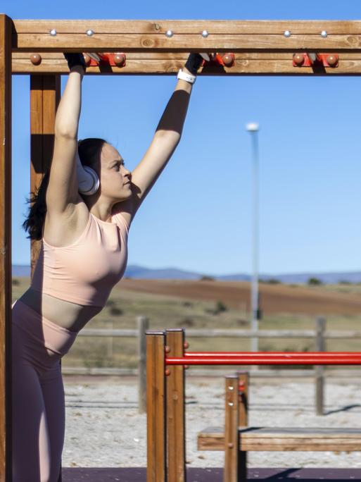 Porträt einer sportlichen Frau beim Klimmzug am Barren an einem sonnigen Tag mit Kopfhörern und Sportkleidung