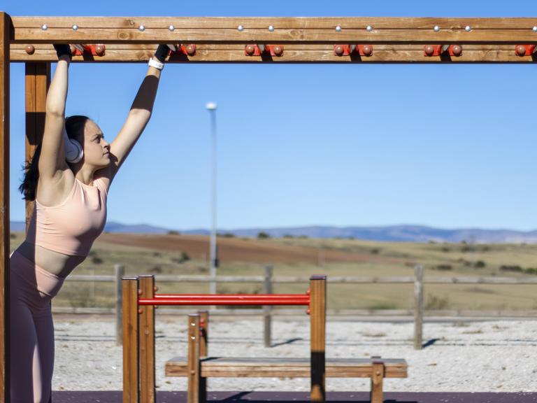 Porträt einer sportlichen Frau beim Klimmzug am Barren an einem sonnigen Tag mit Kopfhörern und Sportkleidung