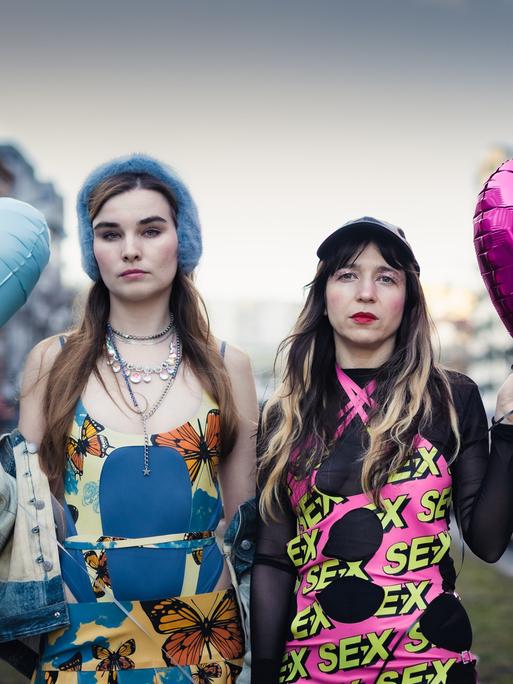 Pauline Schmiechen und Paulita Pappel stehen zwischen zwei Straßen in Berlin, sie tragen bunte Kleidung und einen blauen und einen pinken herzförmigen Luftballon