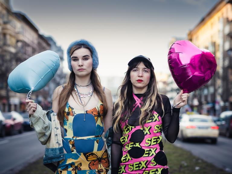 Pauline Schmiechen und Paulita Pappel stehen zwischen zwei Straßen in Berlin, sie tragen bunte Kleidung und einen blauen und einen pinken herzförmigen Luftballon