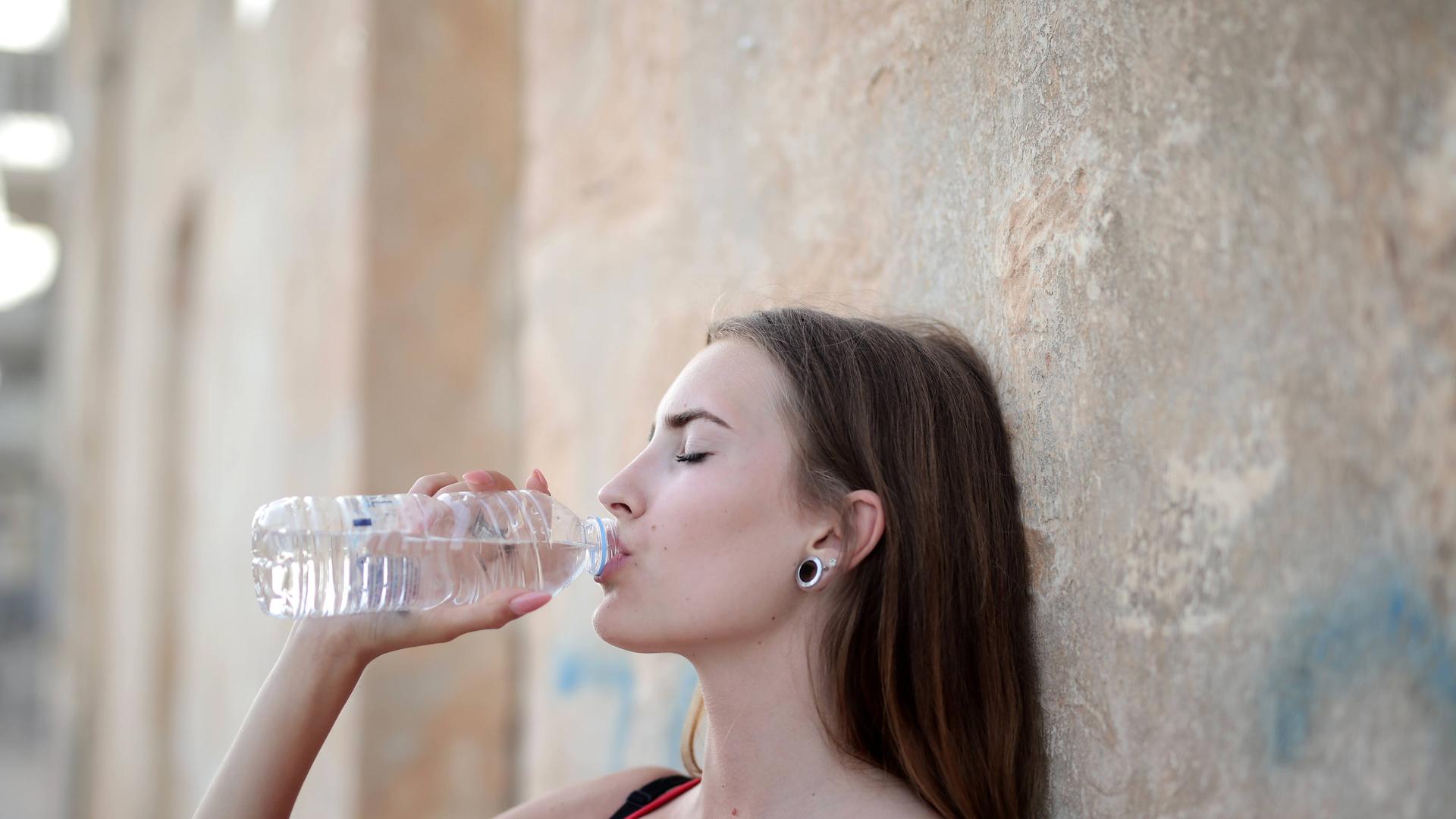 Eine Frau lehnt an einer Mauer und trinkt aus einer Waserflasche.