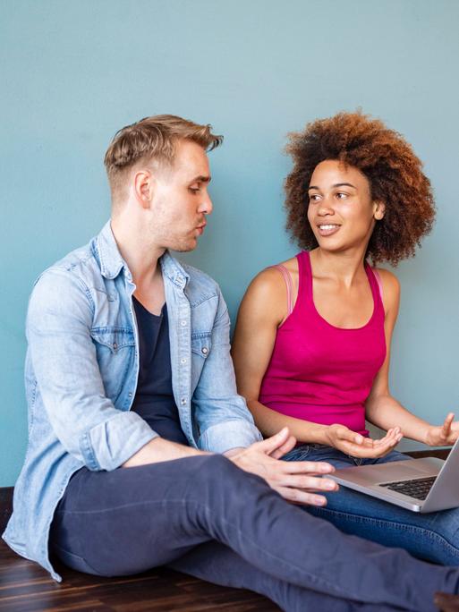 Ein Mann und eine Frau sitzen auf dem Boden. Auf den Beinen der Frau ist ein Laptop. Der Mann erzählt. Ein Mann und eine Frau sitzen auf dem Boden. Auf den Beinen der Frau ist ein Laptop. Der Mann erzählt.