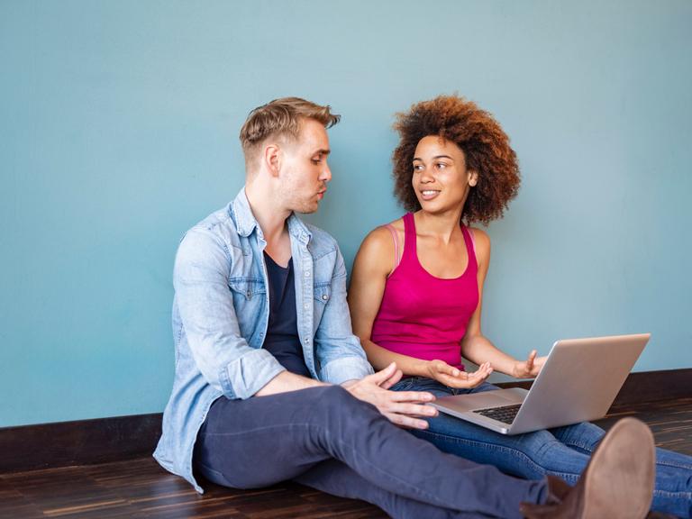 Ein Mann und eine Frau sitzen auf dem Boden. Auf den Beinen der Frau ist ein Laptop. Der Mann erzählt. Ein Mann und eine Frau sitzen auf dem Boden. Auf den Beinen der Frau ist ein Laptop. Der Mann erzählt.