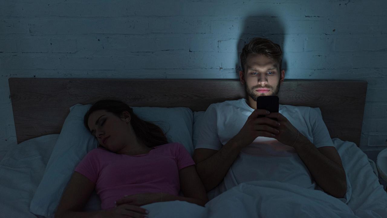 Ein Pärchen liegt im Bett. Der Mann kann nicht schlafen und schaut auf sein Handy. Das Smartphone-Bildschirmlicht hält ihn weiter vom Schlafen ab.