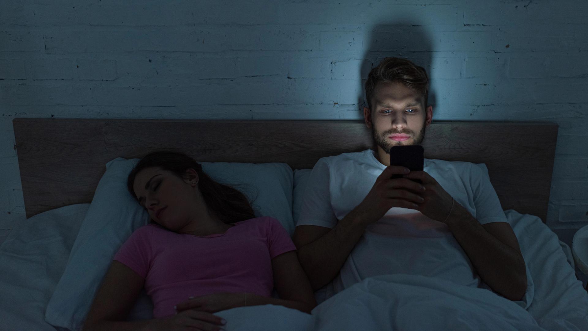 Ein Pärchen liegt im Bett. Der Mann kann nicht schlafen und schaut auf sein Handy. Das Smartphone-Bildschirmlicht hält ihn weiter vom Schlafen ab.