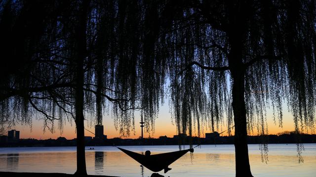 Ein Mensch liegt an einem See in einer Hängematte. Im Hintergrund sind Gebäude erkennbar. Die Sonne geht gerade unter.