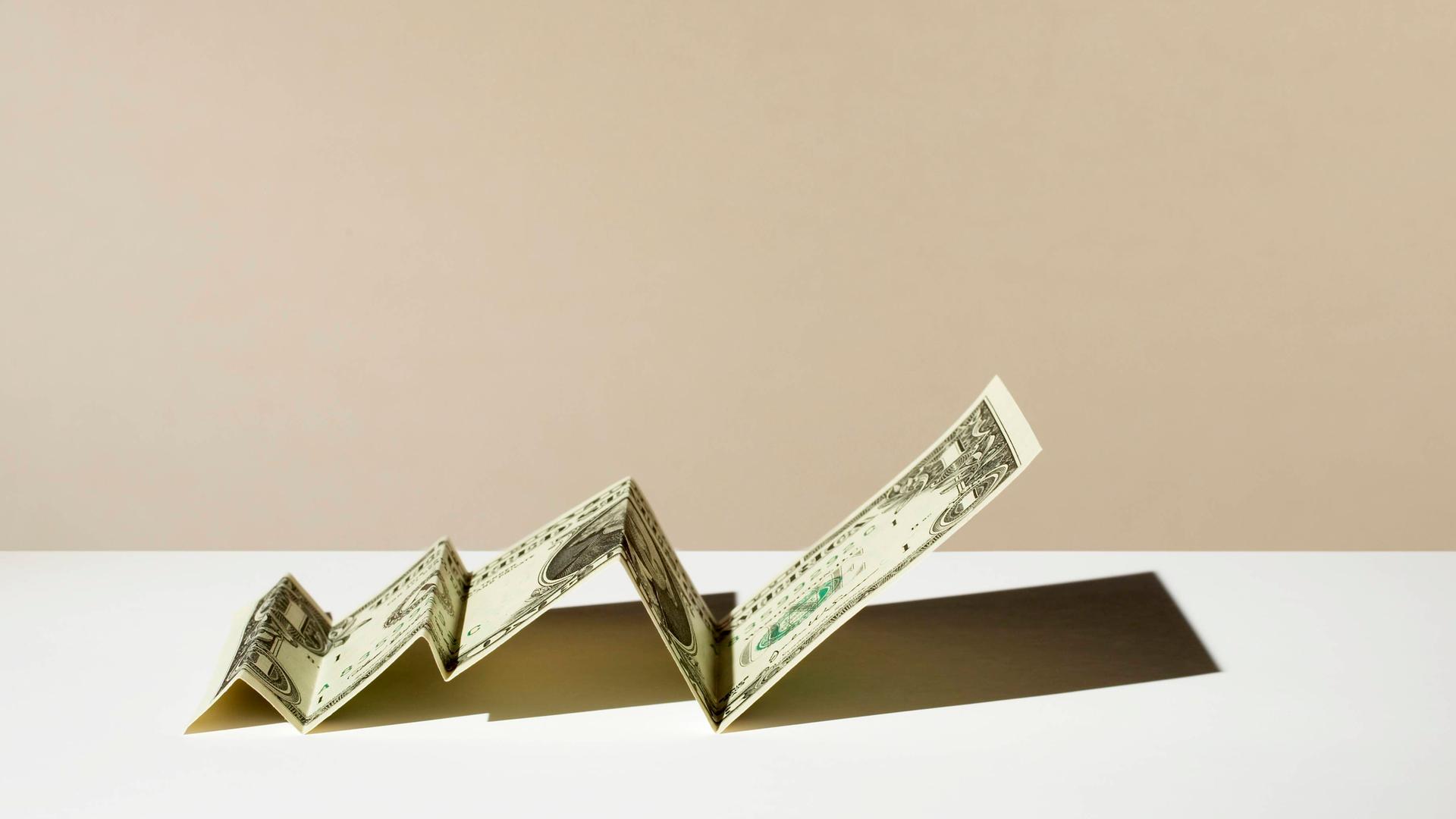 Ein gefalteter Dollar-Schein liegt auf einer Oberfläche. Der Dollar-Schein erinnert durch das Falten an einen Graphen, der ansteigt. Ein gefalteter Dollar-Schein liegt auf einer Oberfläche. Der Dollar-Schein erinnert durch das Falten an einen Graphen, der ansteigt.