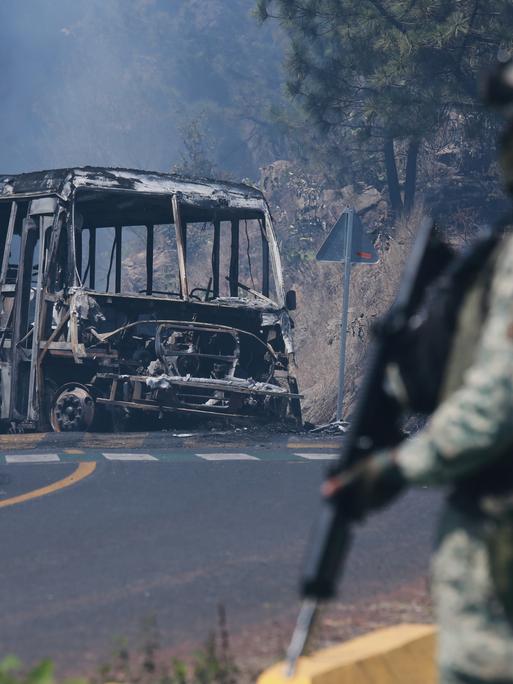 Auf einer Straße in Mexiko liegt ein ausgebrannter Bus. Davor steht ein Soldat, der eine Waffe in der hält.
