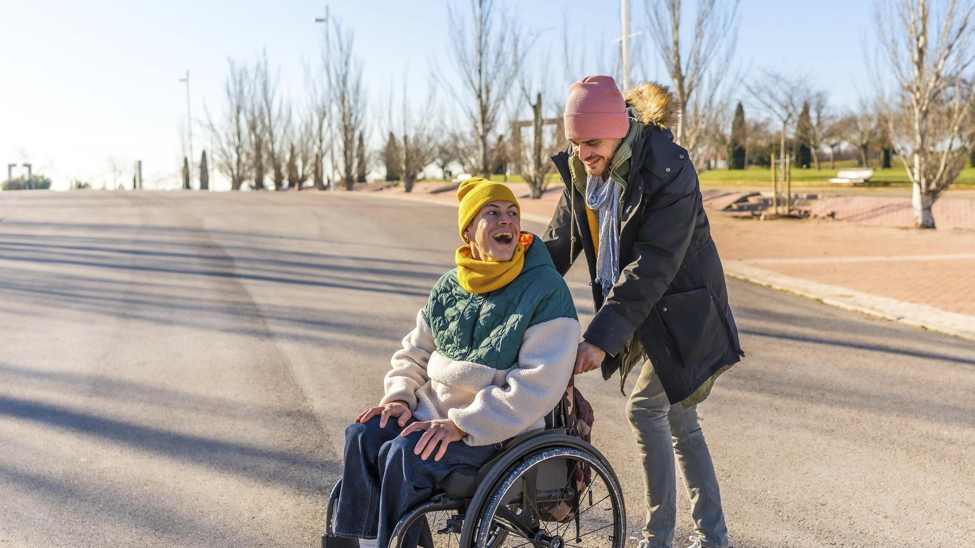 Junger Pfleger hilft einer fröhlichen älteren Frau im Rollstuhl, die einen sonnigen Wintertag in einem Park genießt