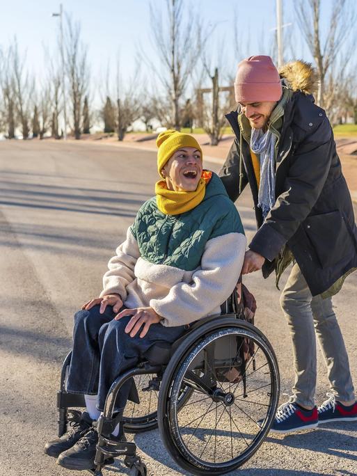 Junger Pfleger hilft einer fröhlichen älteren Frau im Rollstuhl, die einen sonnigen Wintertag in einem Park genießt