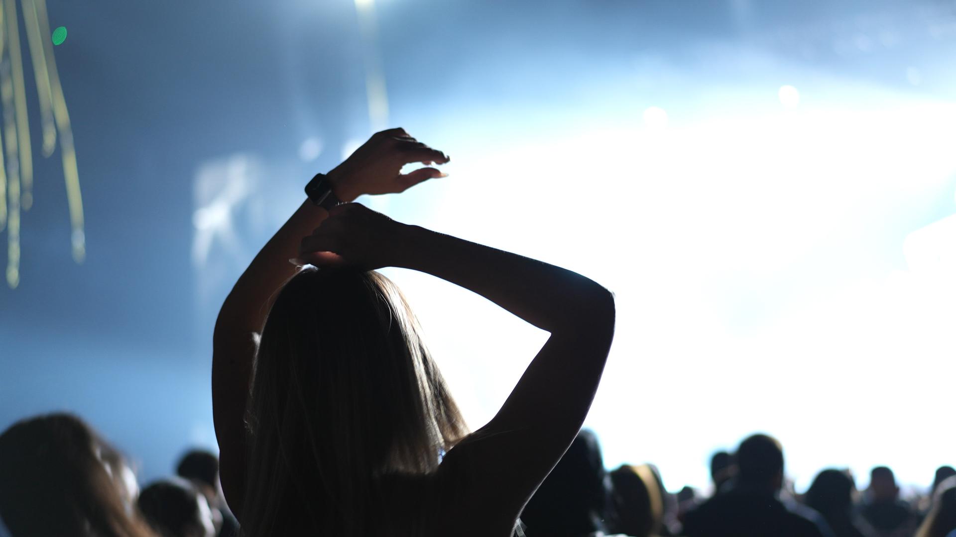 Eine Frau ist bei einem Konzert im Publikum. Sie hält die Arme über ihrem Kopf. Wir sehen sie von hinten. Eine Frau ist bei einem Konzert im Publikum. Sie hält die Arme über ihrem Kopf. Wir sehen sie von hinten.
