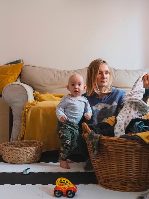 Foto einer jungen Frau, die mit ihrem Kind im Wohnzimmer auf em Boden sitzt und gewaschene Wäsche faltet Foto einer jungen Frau, die mit ihrem Kind im Wohnzimmer auf em Boden sitzt und gewaschene Wäsche faltet