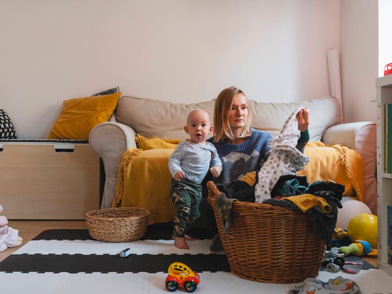 Foto einer jungen Frau, die mit ihrem Kind im Wohnzimmer auf em Boden sitzt und gewaschene Wäsche faltet Foto einer jungen Frau, die mit ihrem Kind im Wohnzimmer auf em Boden sitzt und gewaschene Wäsche faltet