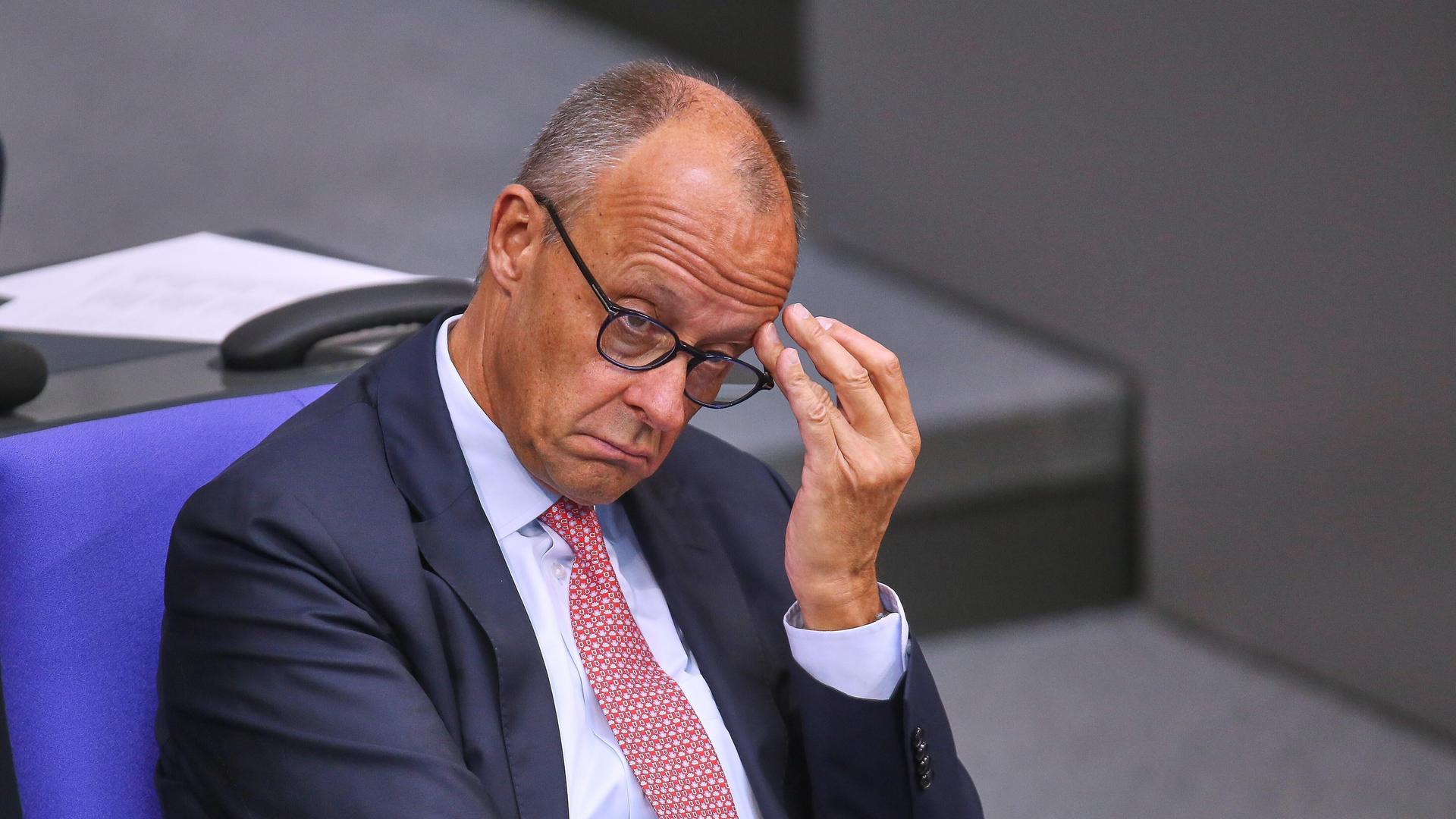 Friedrich Merz sitzt im Bundestag und schaut betreten nach unten. Er fasts sich mit einer Hand an die Brille. 