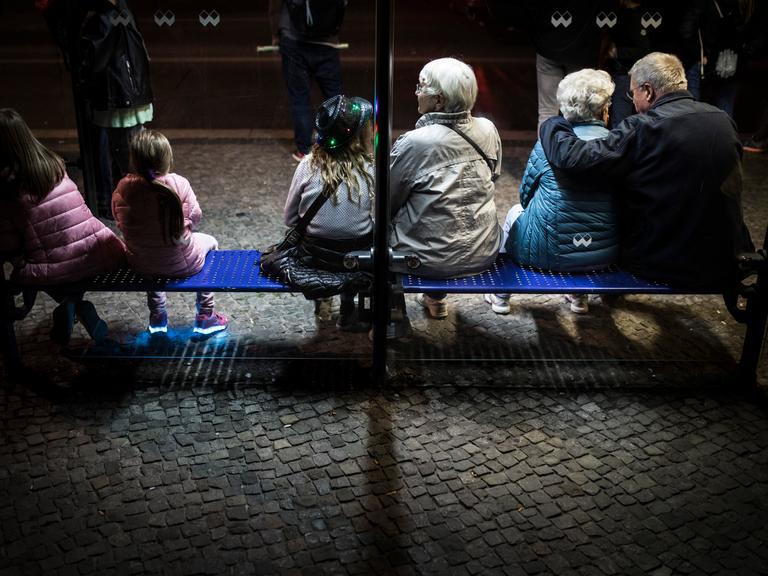 Junge und alte Menschen sitzen im Jahr 2018 auf der Bank einer Bushaltestelle in Berlin