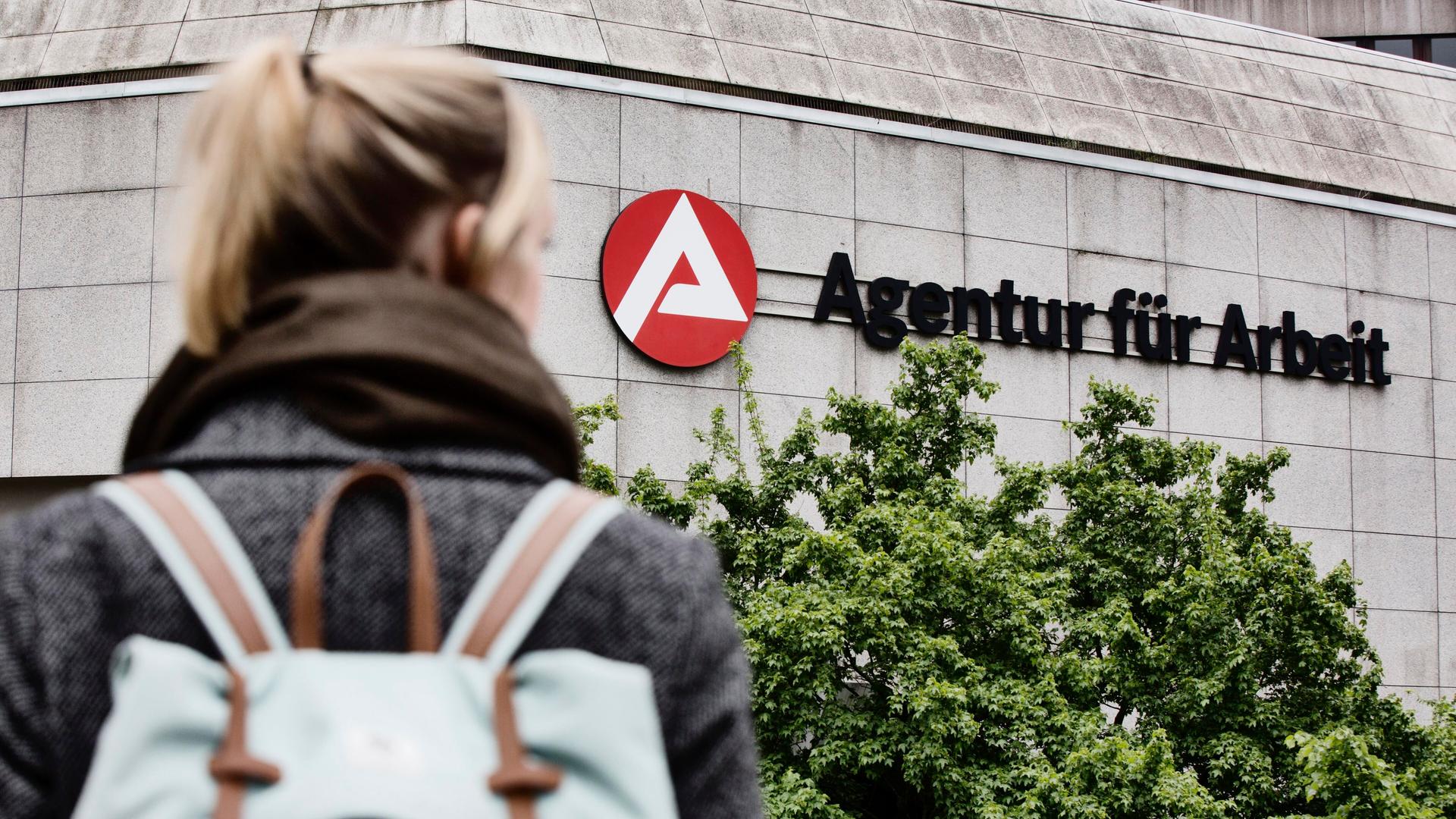 Eine junge Frau steht vor einem Gebäude mit der Aufschrift: "Agentur für Arbeit". Sie steht mit dem Rücken zur Kamera und schaut auf das Gebäude.  