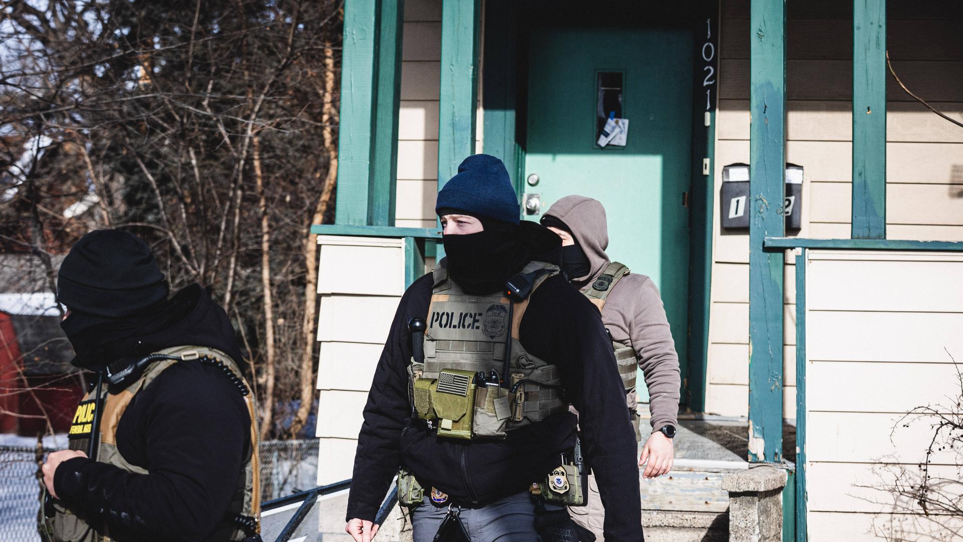 Ein Beamter der US-Einwanderungsbehörde ICE steht vor einem Haus. Er trägt eine Weste mit der Aufschrift "Police" und hat das Gesicht vermummt.