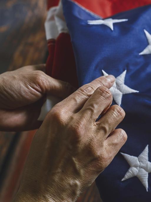 Nahaufnahme von Händen, die ehrfürchtig eine amerikanische Flagge falten und dabei komplizierte Nähte und lebendige Farben betonen. Das Bild vermittelt Respekt, Patriotismus und Liebe zum Detail