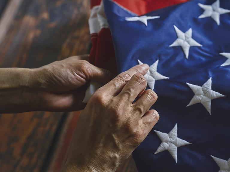 Nahaufnahme von Händen, die ehrfürchtig eine amerikanische Flagge falten und dabei komplizierte Nähte und lebendige Farben betonen. Das Bild vermittelt Respekt, Patriotismus und Liebe zum Detail