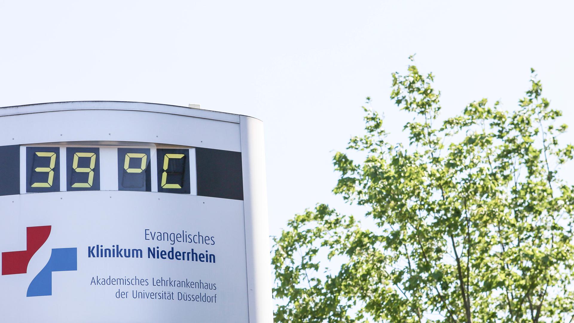 Thermometer vor Krankenhaus zeigt 39 Grad, daneben ein Baum
