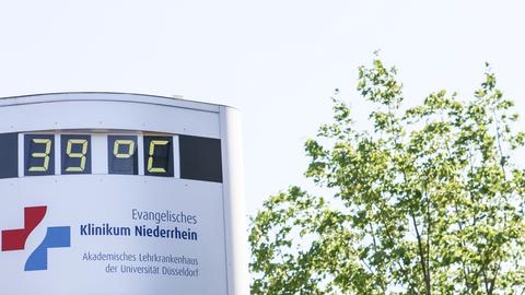 Ein Thermometer vor einem Krankenhaus zeigt 39 Grad, daneben steht ein Baum.