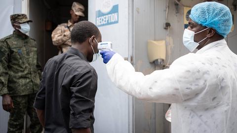 Ein Mediziner misst bei einem Ebola-Screening die Temperatur eines Mannes. Im Hintergrund ist uniformiertes Sicherheitspersonal zu sehen.