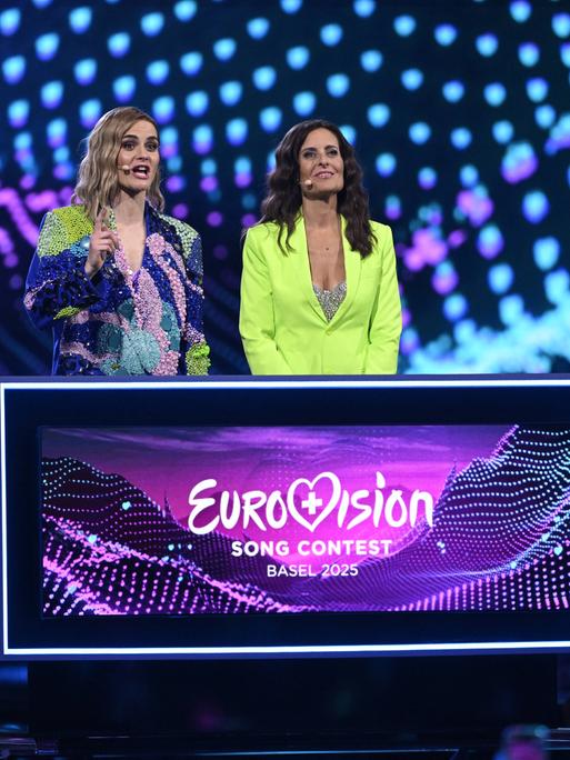 Auf der Bühne des Eurovision Song Contest stehen die beiden Moderatorinnen Hazel Brugger und Sandra Studer. Das Outfit von Brugger hat die Farben der Bühnen: Blau- und Lilatöne. Auf der Bühne des Eurovision Song Contest stehen die beiden Moderatorinnen Hazel Brugger und Sandra Studer. Das Outfit von Brugger hat die Farben der Bühnen: Blau- und Lilatöne.