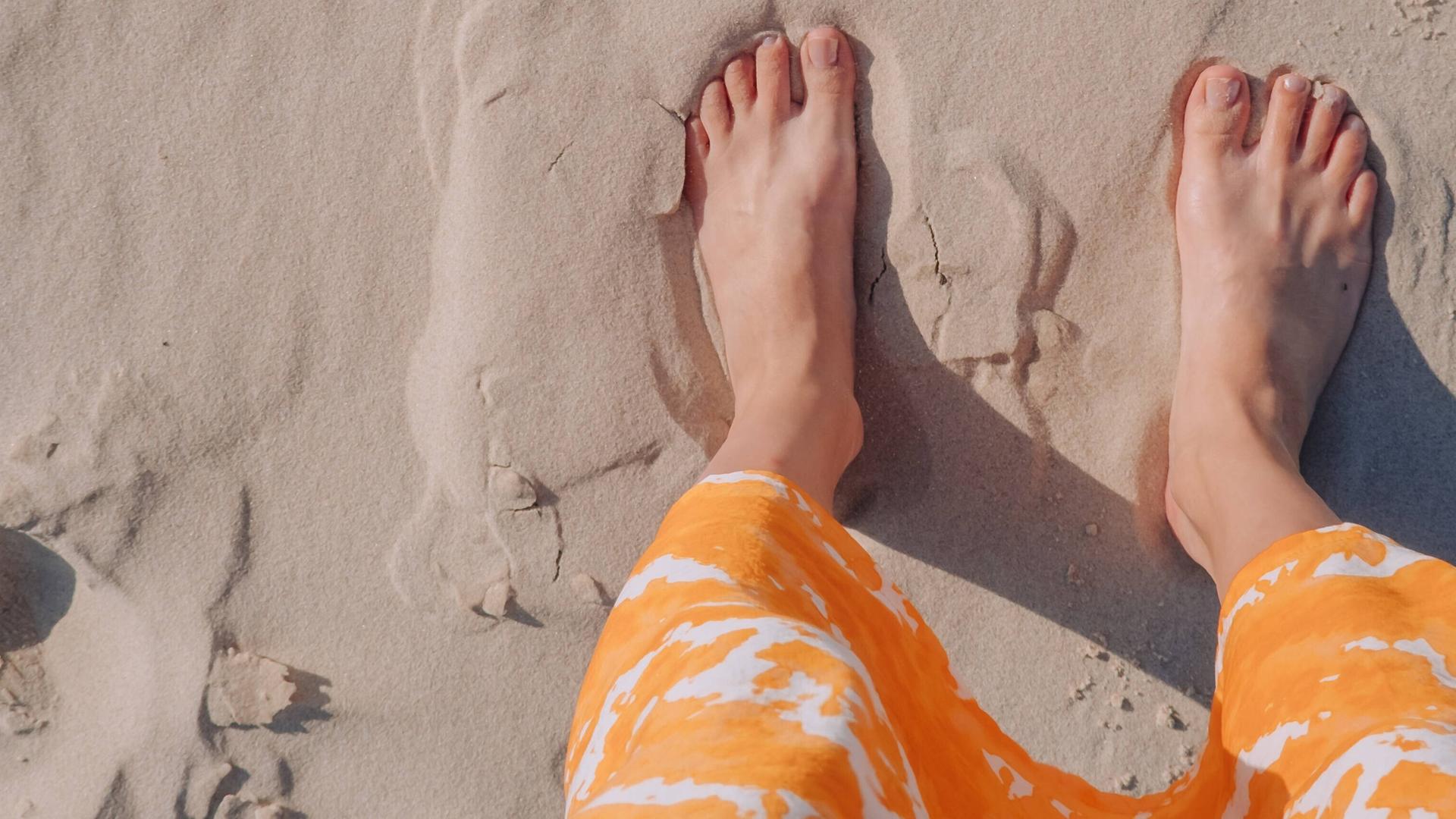 Eine Frau in einem orangefarbenen Rock steht barfuß an einem Strand