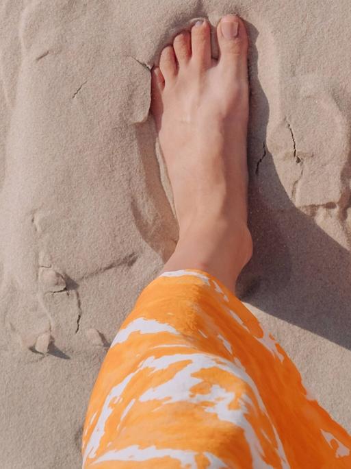 Eine Frau in einem orangefarbenen Rock steht barfuß an einem Strand