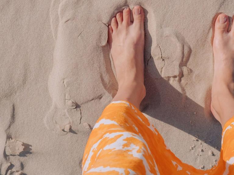Eine Frau in einem orangefarbenen Rock steht barfuß an einem Strand