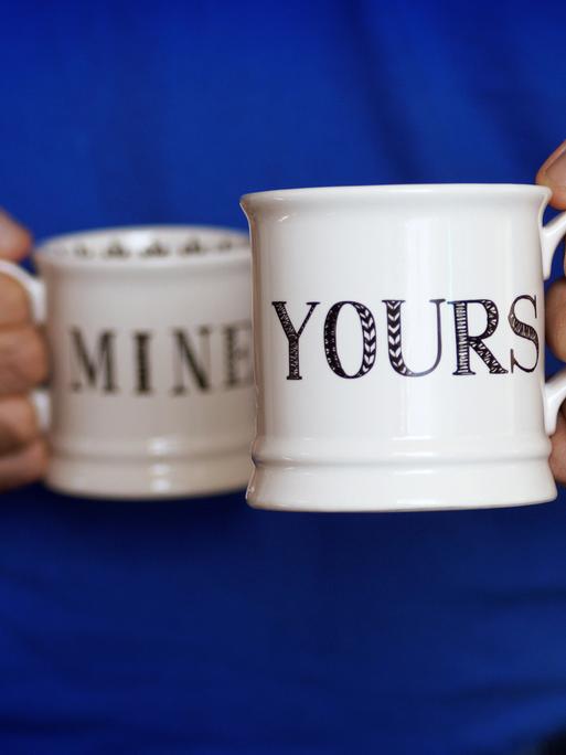 Ein Mann hält zwei Kaffeebecher in seinen Händen: Auf einem steht auf Englisch "mine", auf dem anderen "yours".