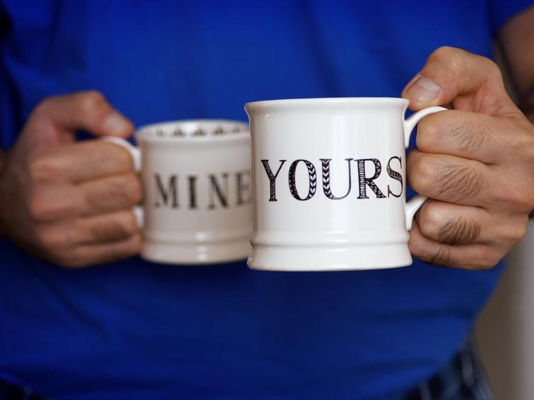 Ein Mann hält zwei Kaffeebecher in seinen Händen: Auf einem steht auf Englisch "mine", auf dem anderen "yours".